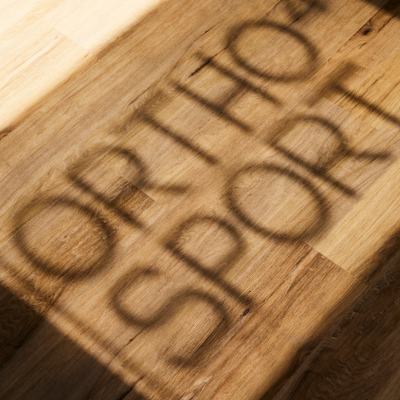 Schatten eines Fensterbeschriftung mit dem Schriftzug ORTHO4SPORT fällt durch Sonnenlicht auf einen Holzparkettboden.